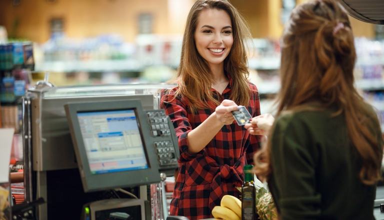 Young lady smiling at and handing her credit card to the cashier who standing behind a counter in a convenience store