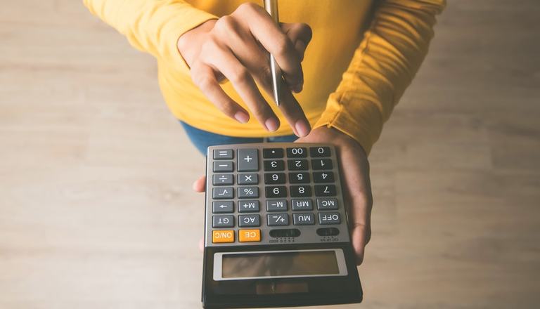 Woman making calculations, pencil in between her fingers in one hand, other hand holding the calculator - calculator is in the middle in foreground, woman's torso and lower body in background