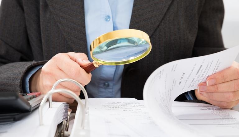 Torso of a suited man conducting an investigation, he holds a magnifying glass in one hand and papers in the other