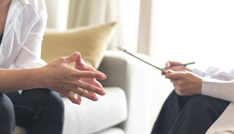 With fingertips pressed together, a patient talks to a therapist, who holds a clipboard