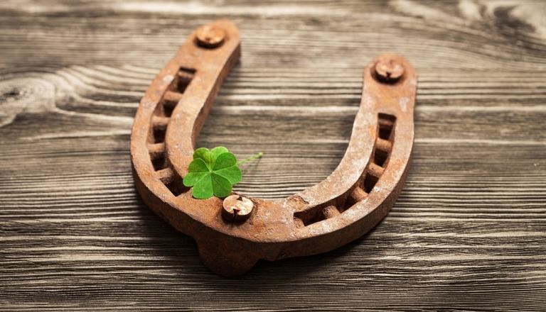 Rusted old horseshoe on a wooden table, with a four-leaf clover on top of it to symbolise good luck