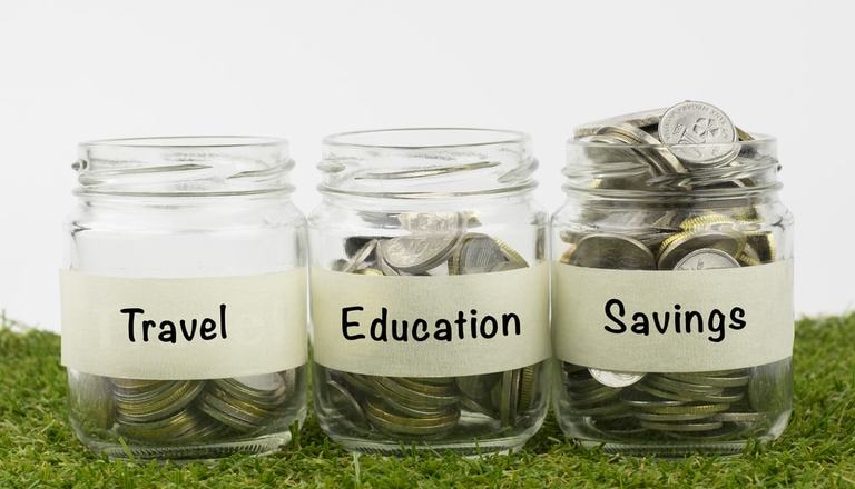 Three glass jars in a row with various amounts of coins in. One jar is labelled "Travel", another is labelled "Education", the third is labelled "Savings"