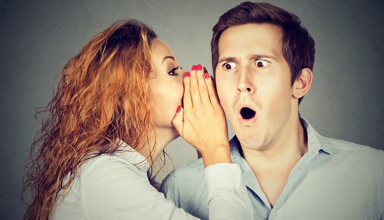 Woman covering mouth with hand whispering to a man who has a shocked expression