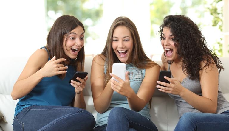 Three women sat on a couch holding cell phones, all are looking at the middle woman's phone and smiling and celebrating