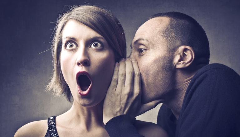 Man telling a woman a secret, cupping his hand against her ear, she has a shocked expression
