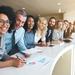Lineup of multi-cultural employees leaning over a desk and smiling at the camera Thumbnail