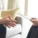 With fingertips pressed together, a patient talks to a therapist, who holds a clipboard Thumbnail